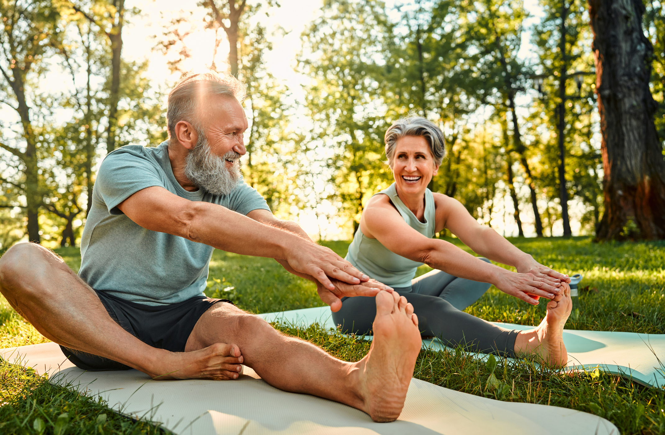 An older couple doing stretching exercises for seniors on yoga mats in a park.