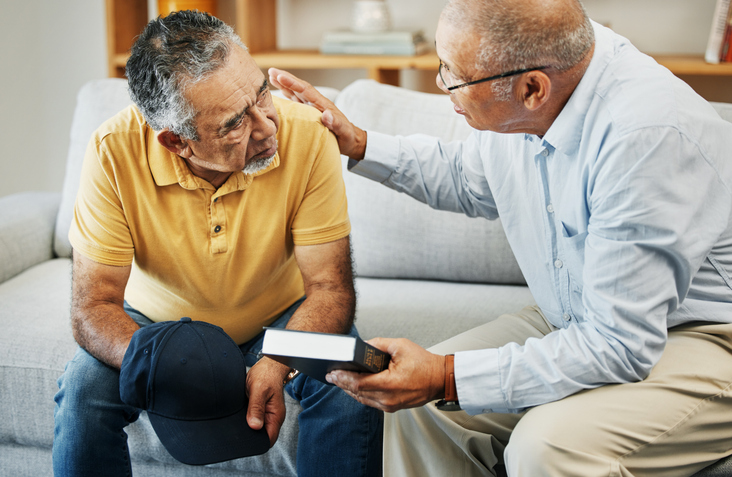 Senior man comforting another while holding a Bible on a living room couch