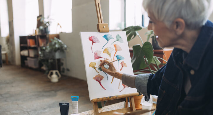 Unrecognizable senior woman painting an art canvas while sitting on a couch in a living room.