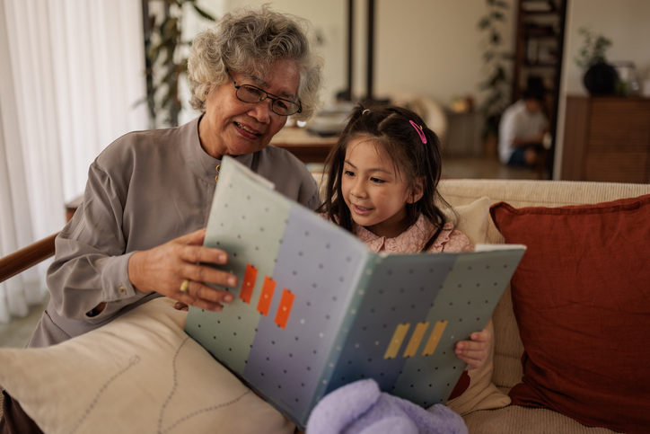 Senior woman smiling at a young girl reading a large book