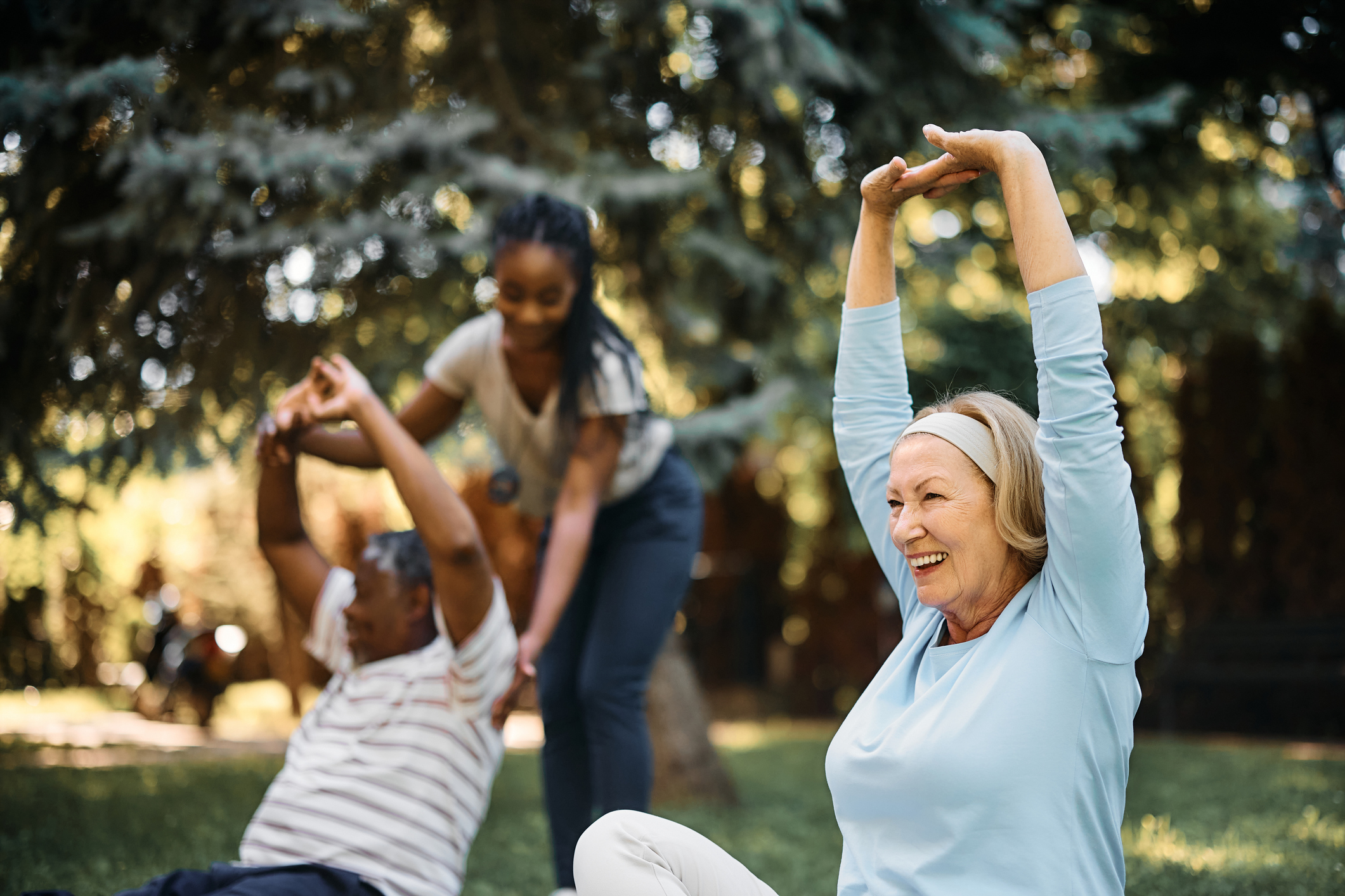 Senior people exercising with physical therapist in the park of a nursing home. Focus is on woman stretching her arms.
