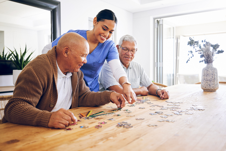 Caregiver assisting two senior men with a jigsaw puzzle at a table