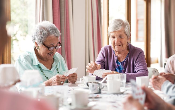 Two senior women playing cards at a table with other seniors