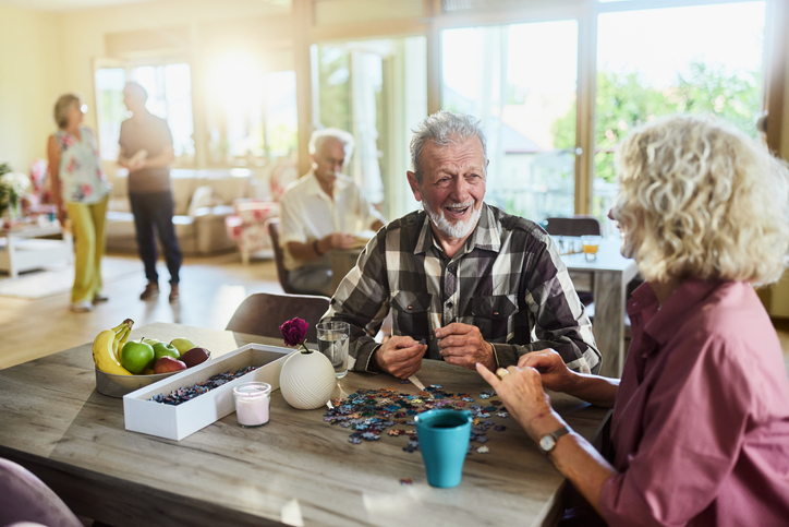 Happy senior man talking to his wife while making jigsaw puzzle in common room at retirement home.