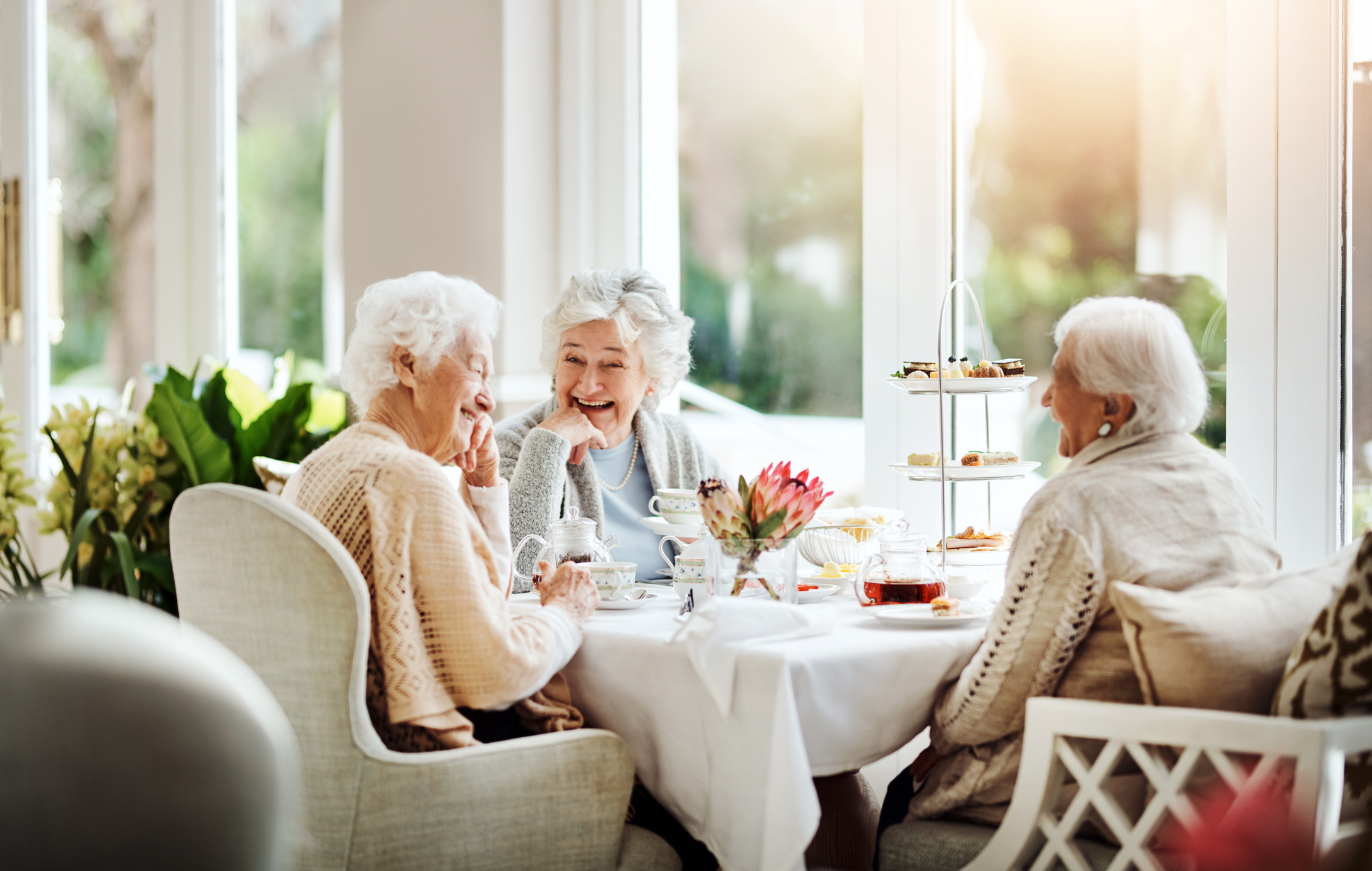 Three senior women laughing at a table with tea and treats
