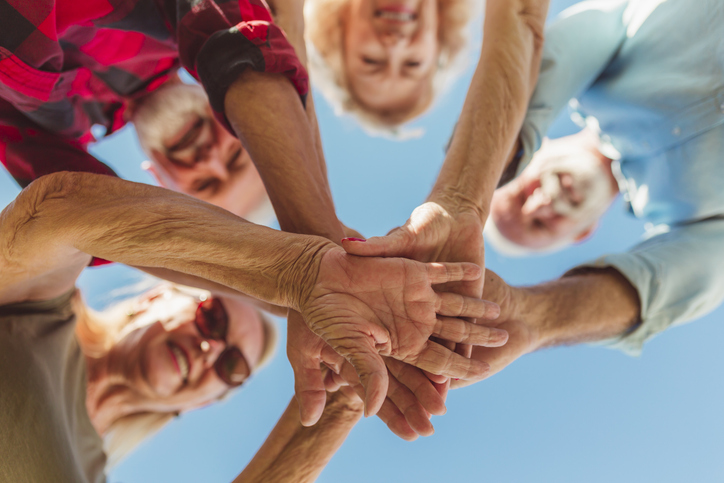 Low angle view of group of cheerful active senior people enjoying sunny summer day outdoors, gathered in circle, holding hands all together in the middle