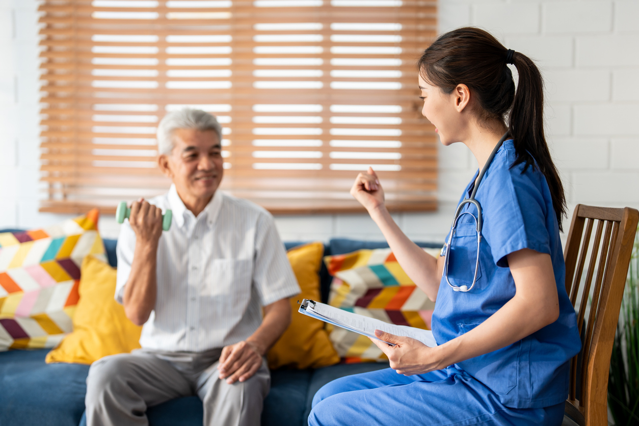 Asian senior elderly man doing physiotherapist with support from nurse. Mature older male patient sit on sofa in living room, using dumbbell exercise with young caregiver at home health nursing care.