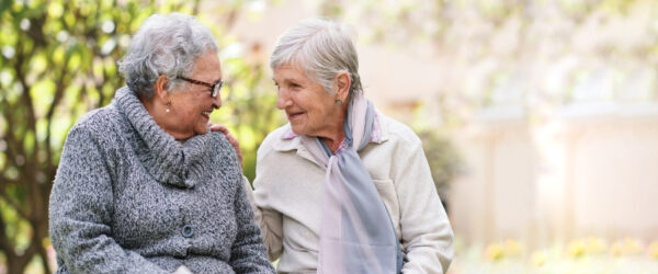 Two older women sitting outdoors, smiling warmly at each other and sharing a friendly moment.