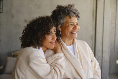 Smiling older woman with gray hair embraced by a younger woman, both sharing a warm and affectionate moment.