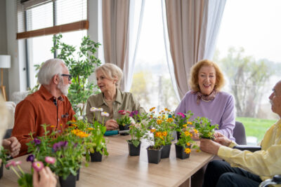 Group of older adults sitting at a table, smiling and enjoying a gardening activity with potted flowers.