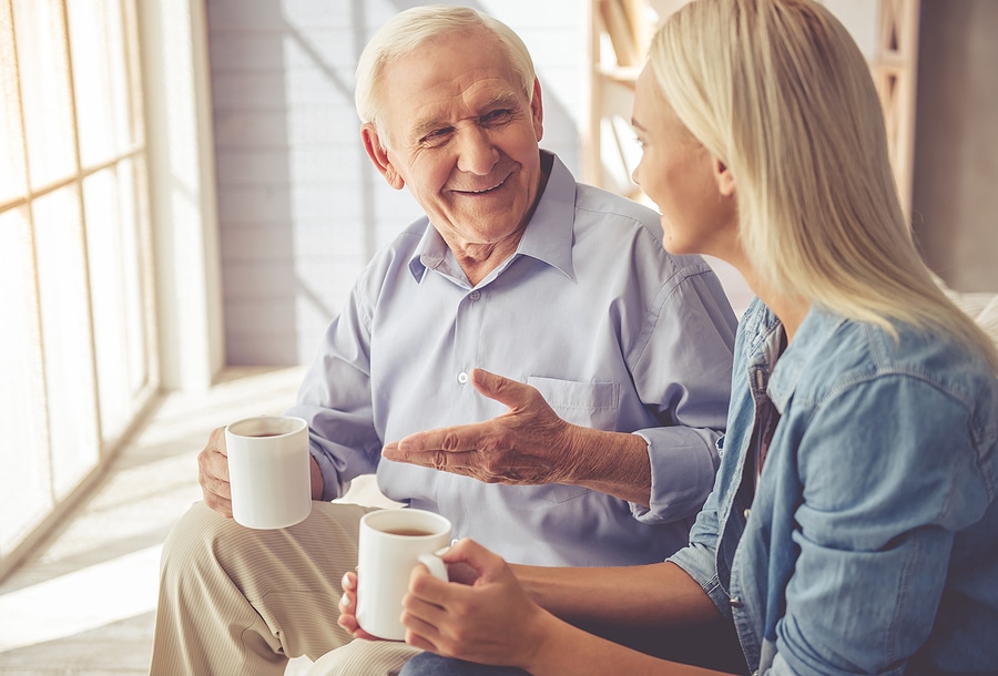 Senior father and adult daughter sitting together on a couch, sharing tea and conversation about Medicaid assisted living waiver.